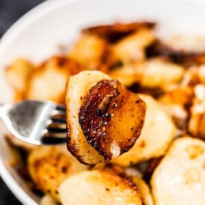 Close-up of a fork holding a slice of golden-brown roasted potato, showcasing the delicious allure of this pan-fried potatoes recipe, with a bowl filled with similarly crispy, seasoned delights in the background.