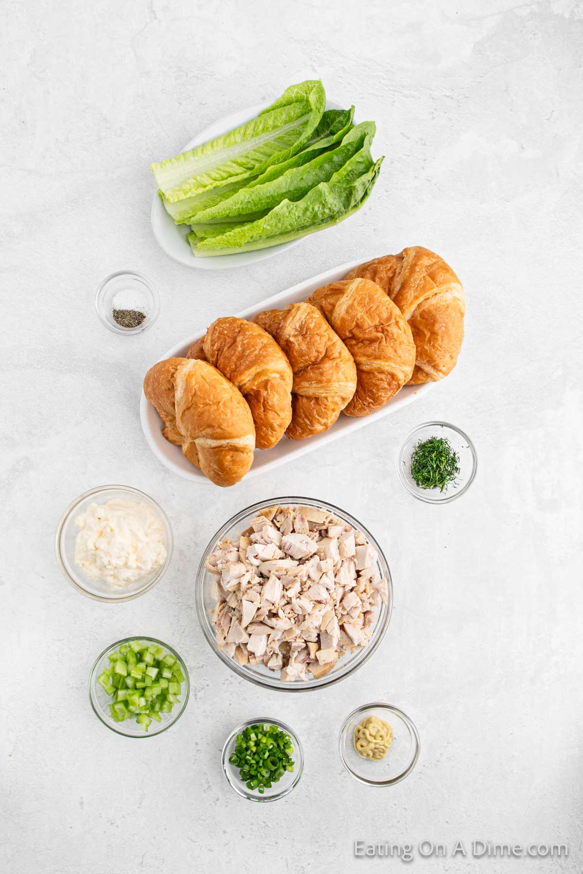 Ingredients for making the best chicken salad recipe croissant sandwiches are laid out on a light surface. Items include chopped chicken, croissants, leafy greens, diced celery, chopped green onions, mayonnaise, mustard, dill, and black pepper.