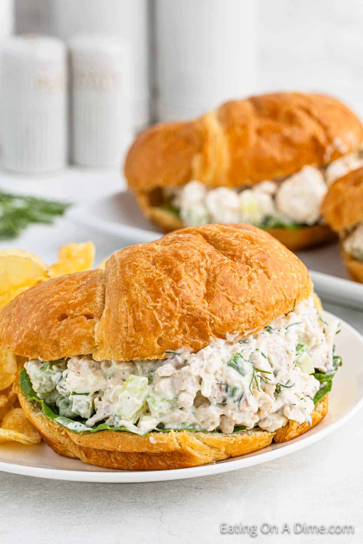 A close-up of a croissant sandwich filled with a creamy chicken salad mixture, garnished with green leafy lettuce. Another similar sandwich is blurred in the background, and there are potato chips on the side. The text "Eating on a Dime.com" is visible at the bottom, showcasing the best chicken salad recipe.