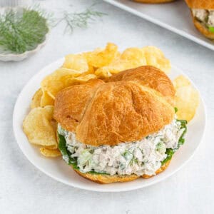 A croissant sandwich filled with the best chicken salad recipe and leafy greens is served on a white plate. The sandwich is accompanied by a side of potato chips, and a small sprig of fresh dill is visible in the background. The setting is light and airy.