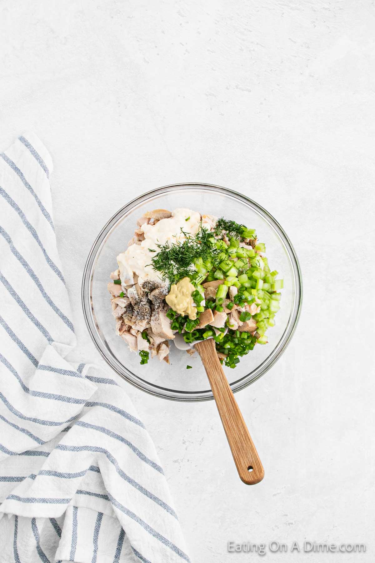 A clear bowl filled with mayo, shredded chicken, chopped celery, herbs, and seasoning sits on a white surface. A wooden spoon rests inside the bowl, while a blue and white striped cloth is folded beside it—truly the best chicken salad recipe in the making.