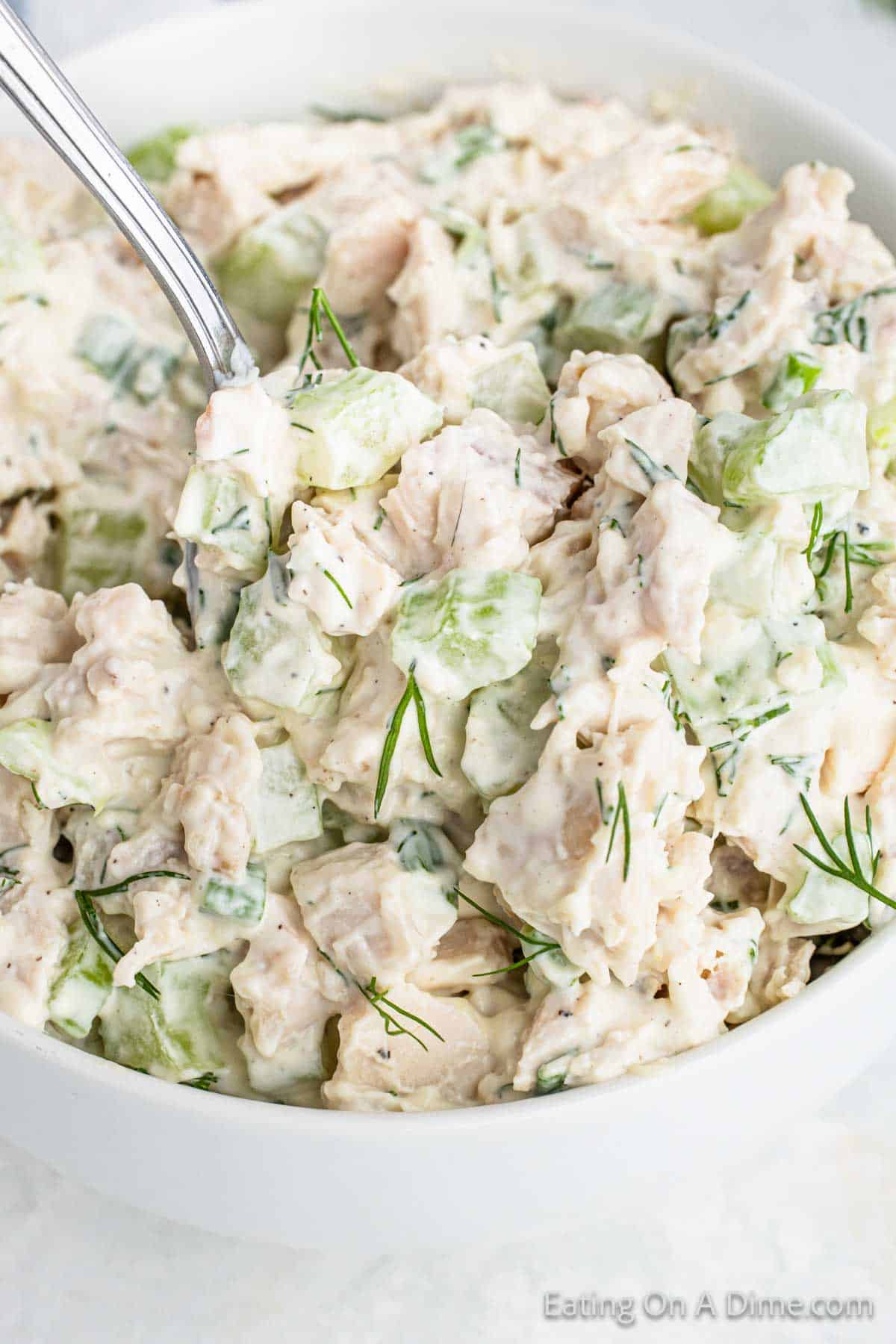 A white bowl filled with a creamy chicken salad, featuring diced chicken, celery, and fresh dill. A silver spoon is resting in the salad, which looks finely mixed and ready to serve—truly the best chicken salad recipe you'll ever find.