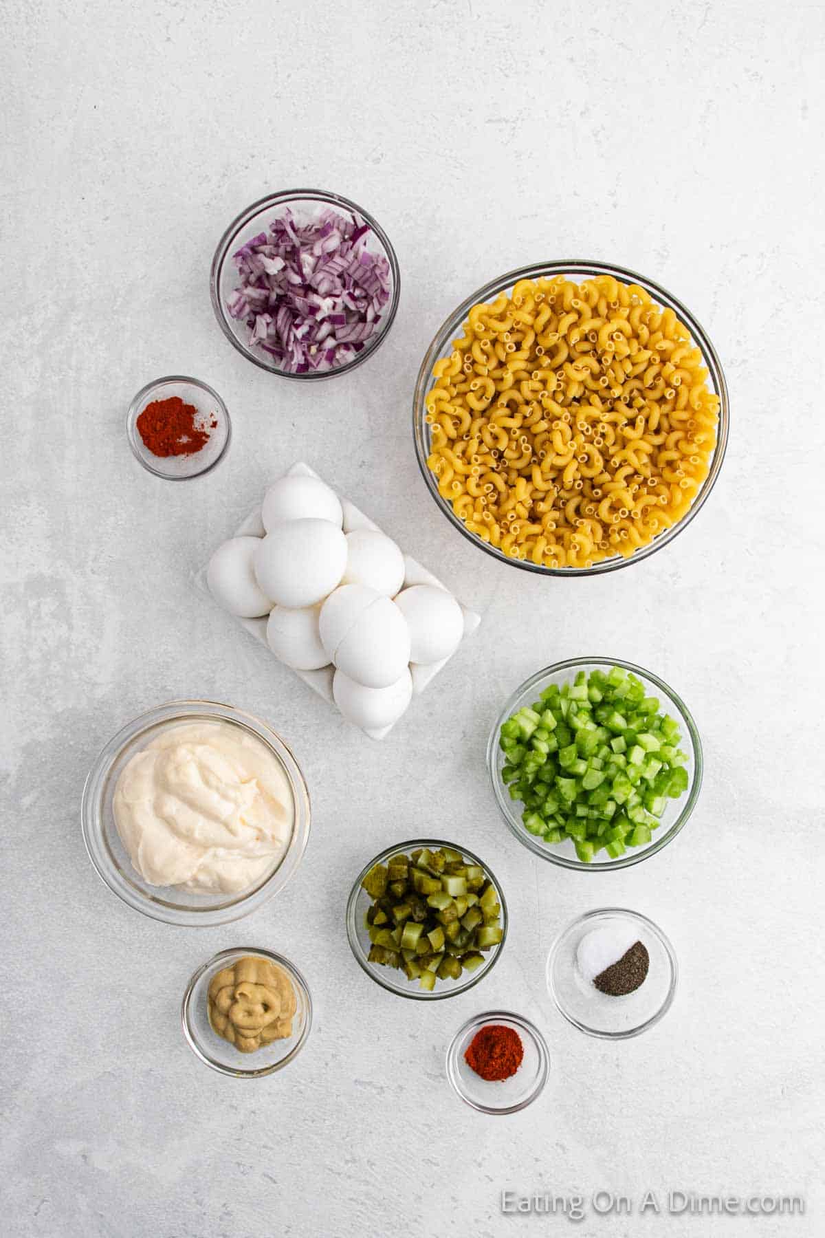 Ingredients for a deviled egg macaroni salad arranged on a white surface. Includes bowls of macaroni, diced red onion, hard-boiled eggs, mayonnaise, pickles, chopped green bell pepper, mustard, and small dishes with salt, paprika, and pepper.