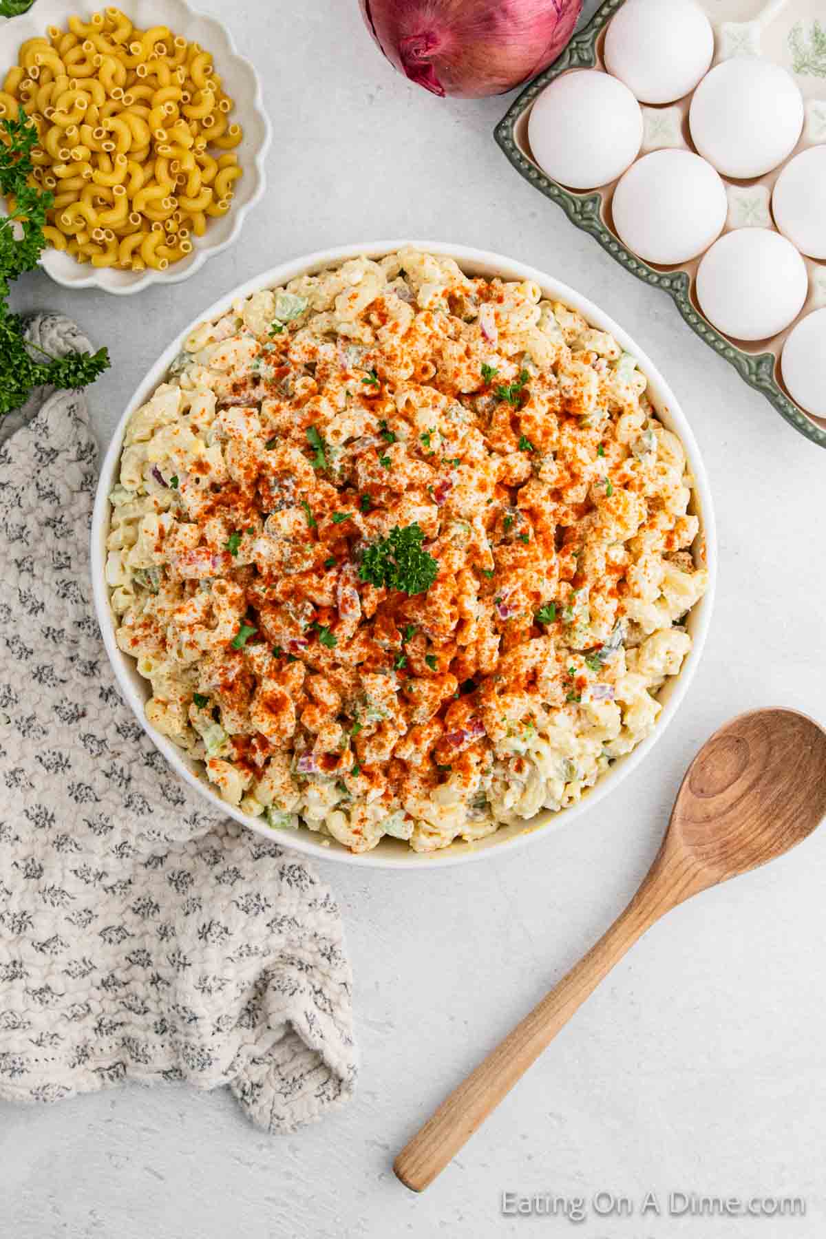 A bowl brimming with Deviled Egg Macaroni Salad is topped with paprika and parsley. Nearby, ingredients like eggs, uncooked macaroni, and an onion await. A wooden spoon and patterned cloth rest beside the bowl on a light-colored surface.