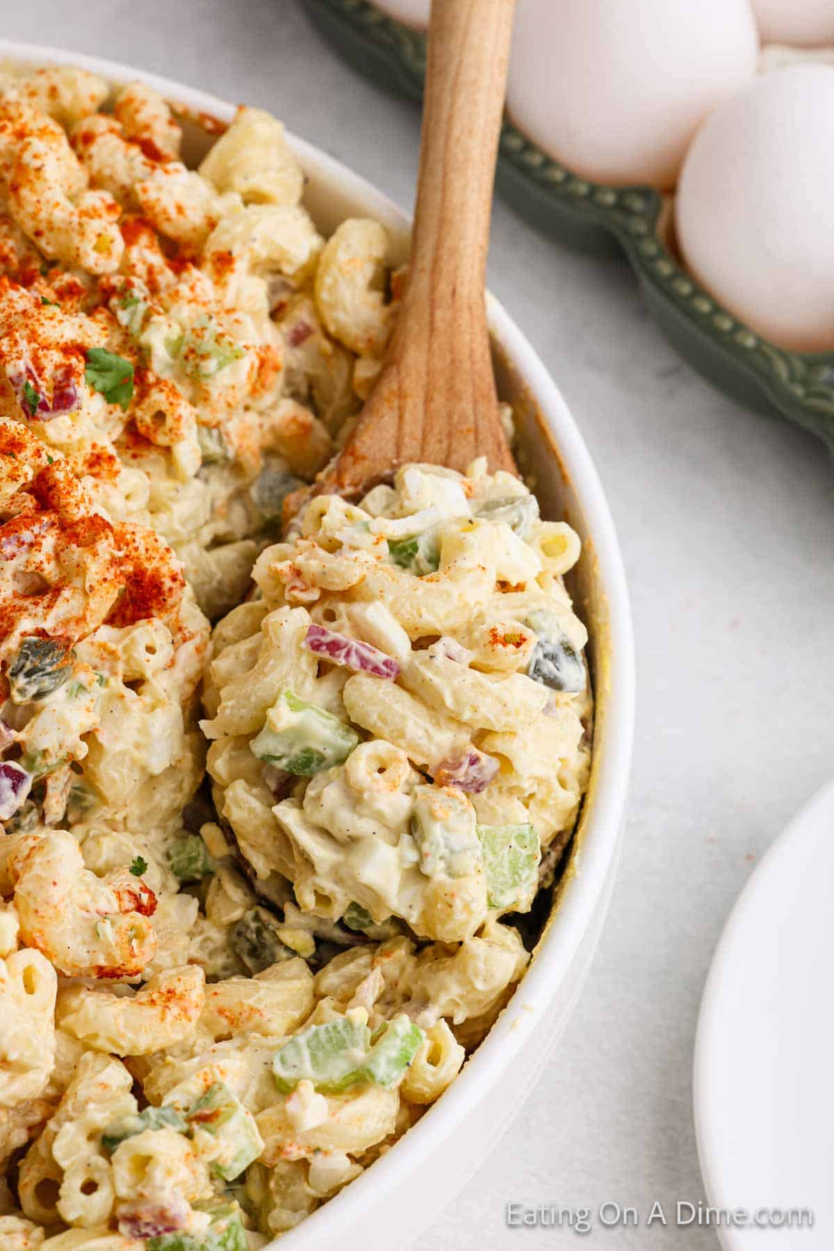 A creamy Deviled Egg Macaroni Salad in a white dish, featuring diced celery, red onion, and green peppers, sprinkled with paprika. A wooden spoon is scooping out a portion. Eggshells are visible in the background.