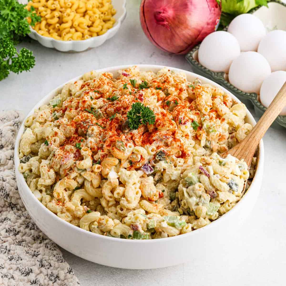 A bowl of creamy deviled egg macaroni salad garnished with paprika and parsley. A wooden spoon is placed in the bowl. Nearby, there are eggs, dry macaroni, a red onion, and fresh parsley on the table.