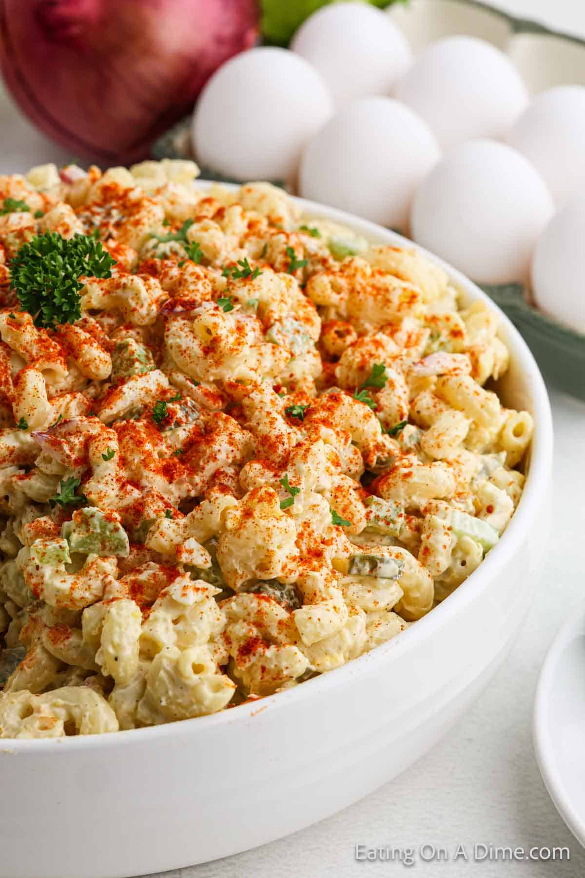 A close-up of a bowl of deviled egg macaroni salad sprinkled with paprika, garnished with parsley. In the background, a few eggs and a red onion are visible.