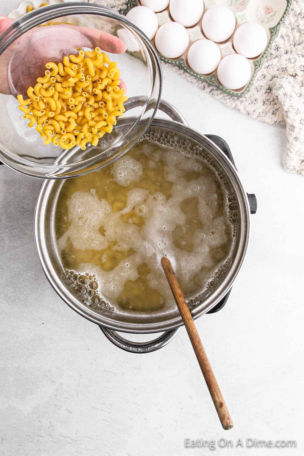 Hand pouring uncooked macaroni into a pot of boiling water on a stove. A wooden spoon rests in the pot, ready for stirring up some deviled egg macaroni salad magic. A carton of eggs and a floral cloth are in the background on a white countertop, setting the scene for this delicious creation.