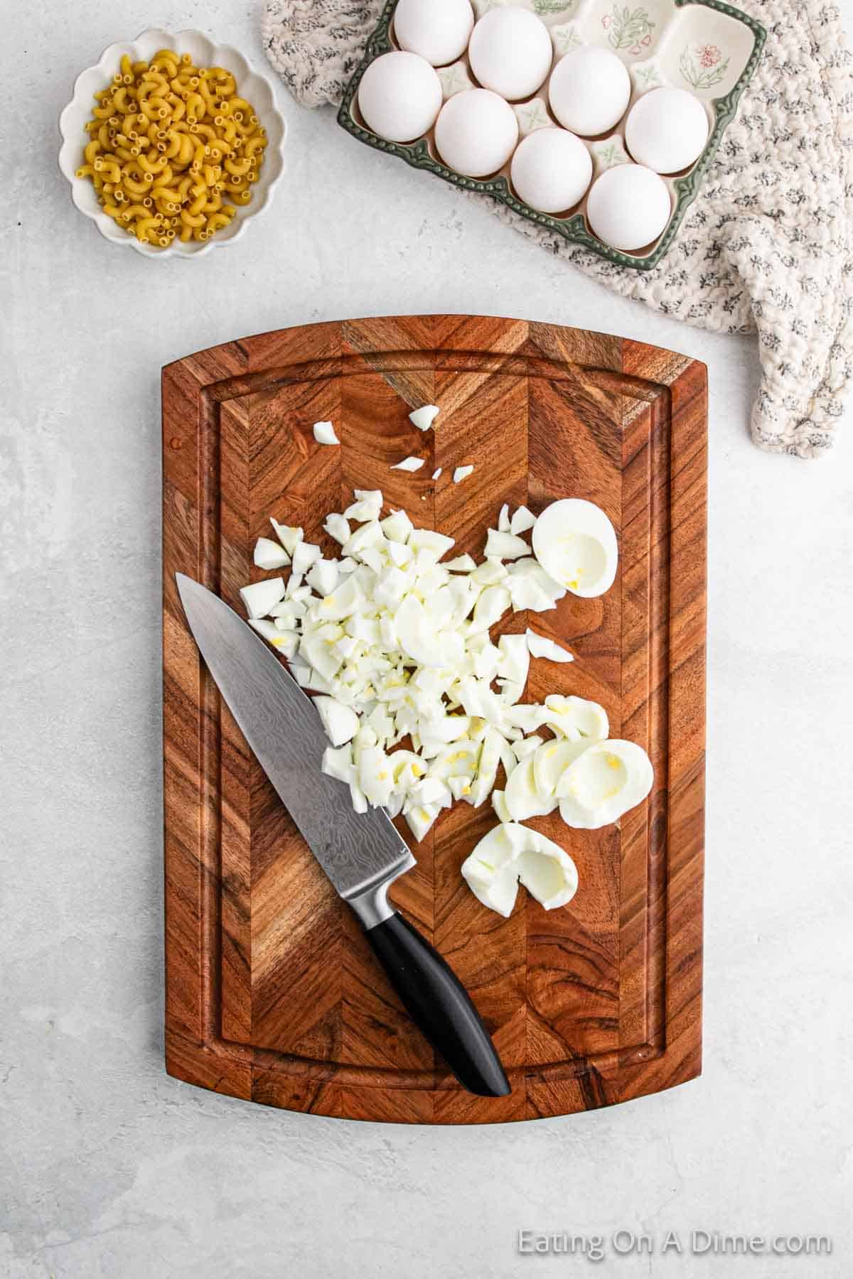 Chopped boiled eggs on a wooden cutting board with a knife, alongside ingredients for deviled egg macaroni salad—a bowl of uncooked macaroni and a carton of eggs—all arranged on a patterned cloth over a light gray surface.