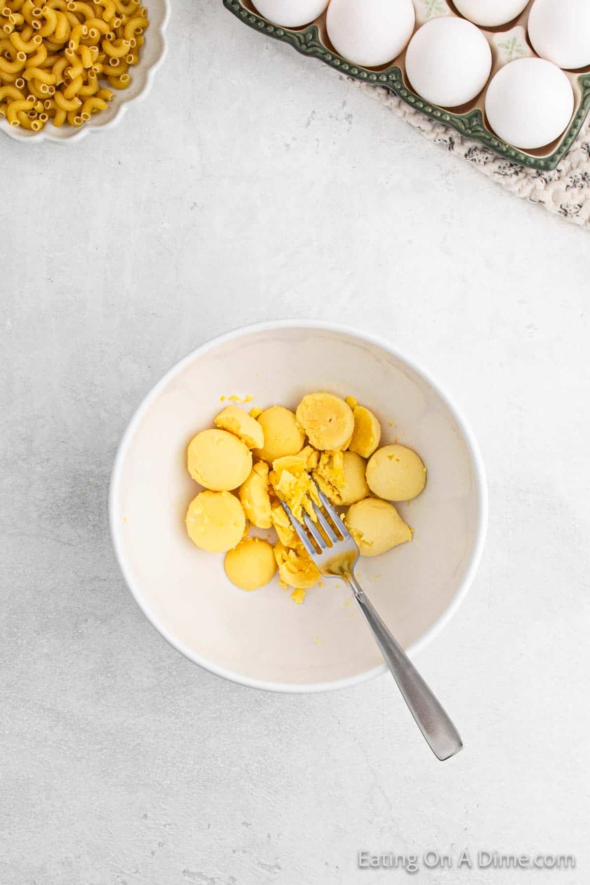 A bowl of sliced boiled egg yolks is being mashed with a fork, ready to transform into a creamy deviled egg macaroni salad. Nearby, there's a plate of macaroni and whole eggs on a patterned cloth, all set against a light gray surface.