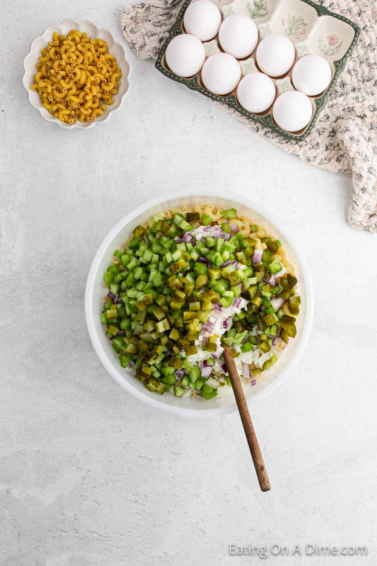 A top view of a kitchen counter with ingredients for Deviled Egg Macaroni Salad. A large bowl contains diced pickles, celery, and onions with a wooden spoon. Nearby are a bowl of uncooked elbow macaroni and a tray of eggs on a floral cloth.