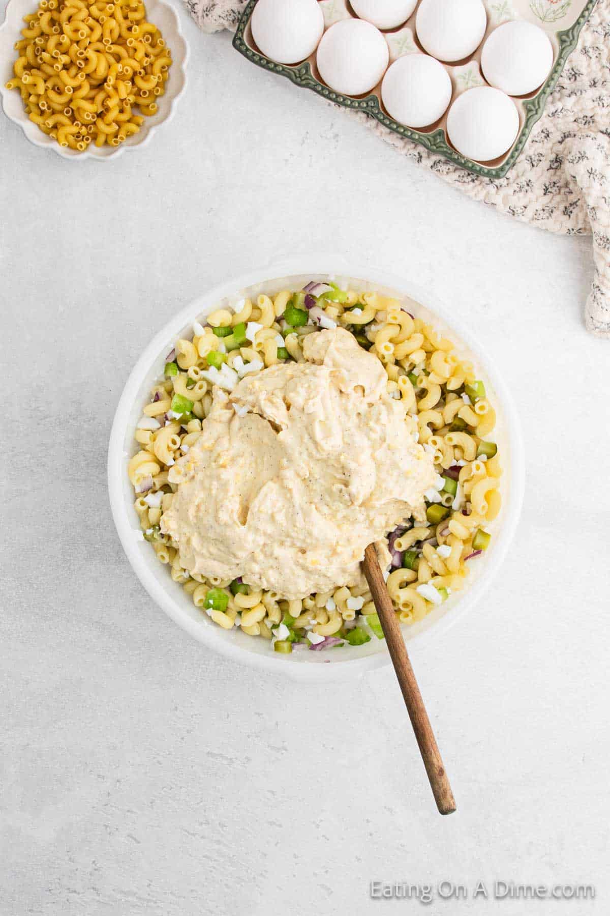 A bowl of deviled egg macaroni salad, bursting with chopped vegetables and topped with creamy dressing, sits alongside a wooden spoon. Nearby, a dish of dry macaroni, a carton of eggs, and a floral cloth rest on the white surface.