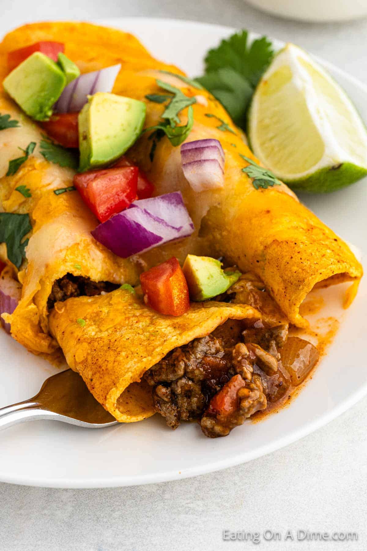 A plate with two Easy Ground Beef Enchiladas topped with melted cheese, diced avocado, tomato, red onion, and cilantro. A lime wedge is placed beside the enchiladas, and a fork is cutting into one of them, revealing the seasoned beef and vegetables inside.
