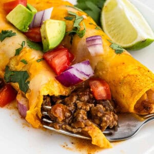 Close-up of easy ground beef enchiladas topped with diced avocado, red onion, tomato, and cilantro. A lime wedge sits in the background. A fork cuts into the enchilada revealing the seasoned beef filling. The dish is served on a white plate.