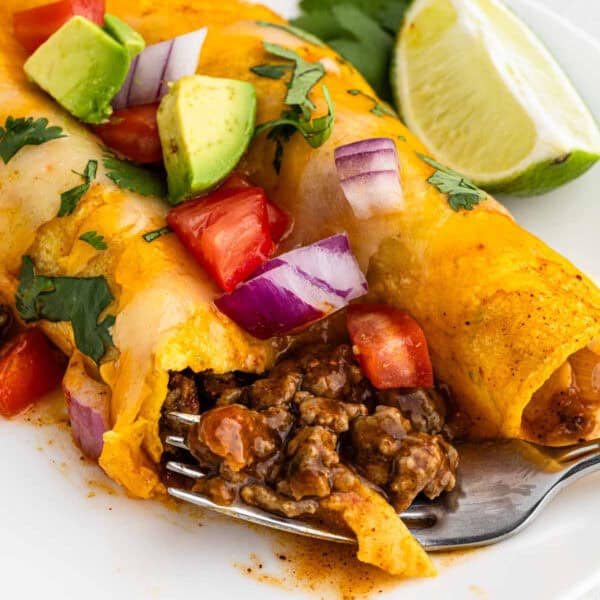 Close-up of easy ground beef enchiladas topped with diced avocado, red onion, tomato, and cilantro. A lime wedge sits in the background. A fork cuts into the enchilada revealing the seasoned beef filling. The dish is served on a white plate.