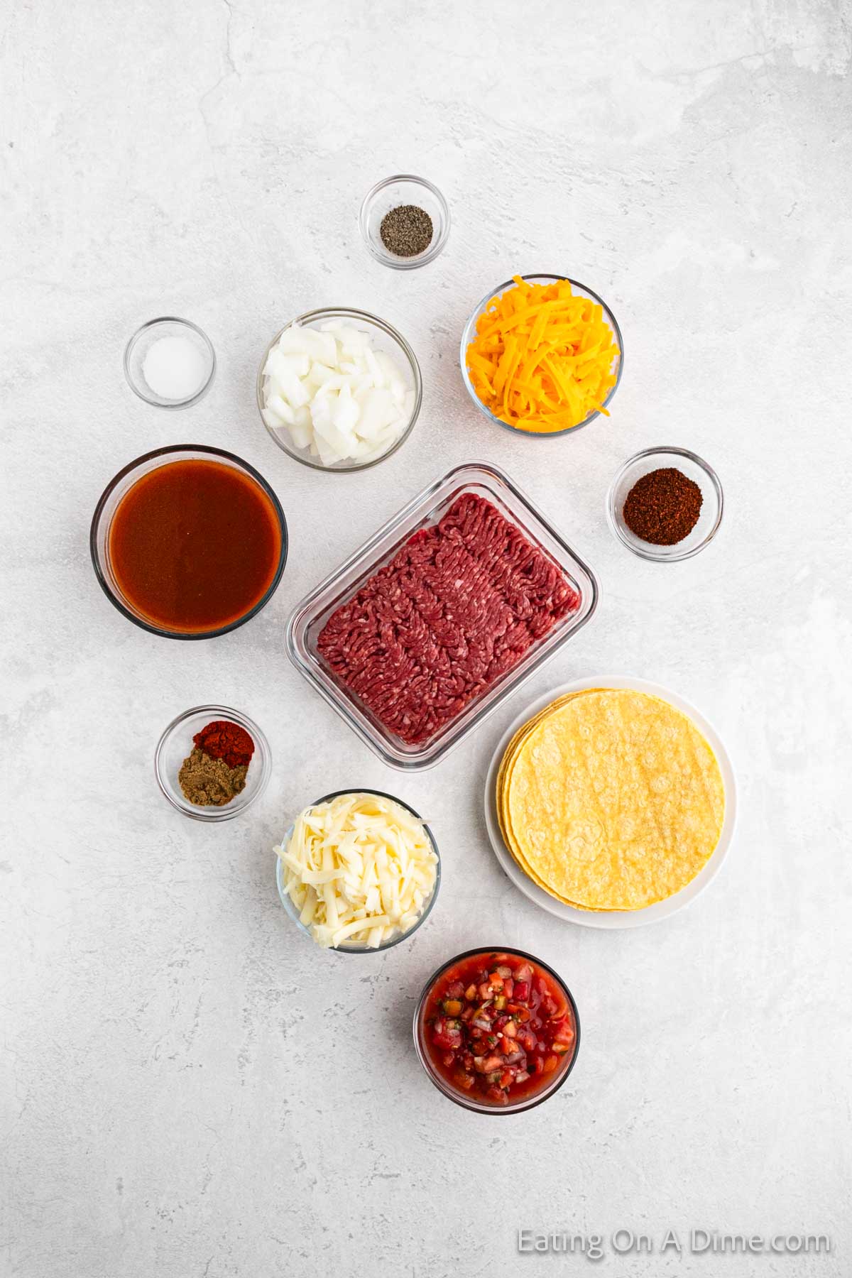 A flat lay photo of various ingredients on a light surface. Central is ground beef in a container, perfect for Easy Ground Beef Enchiladas. Surrounding are small bowls of diced onions, shredded cheddar, tortilla chips, salsa, shredded cheese, spices, and a cup of sauce. Text in the corner reads "Eating On A Dime.com".