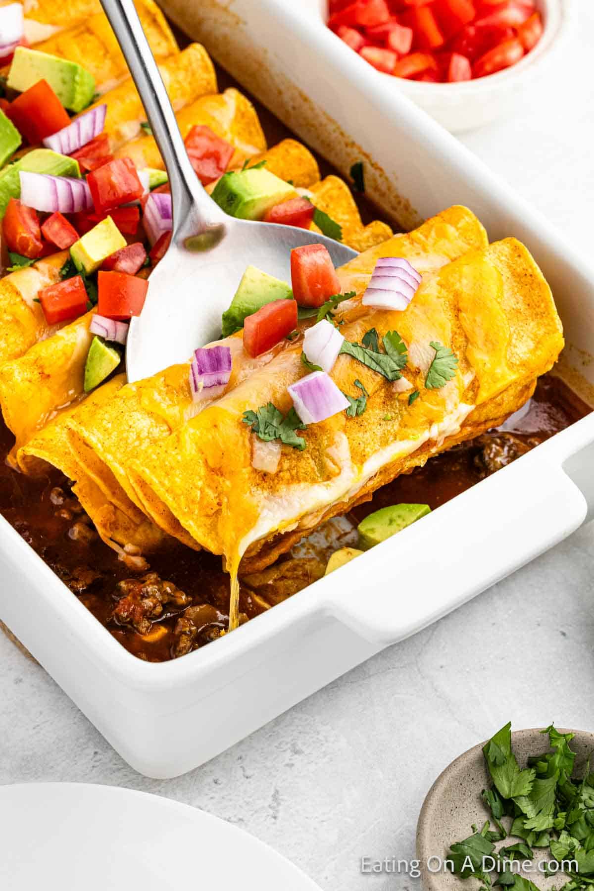 A white rectangular dish filled with Easy Ground Beef Enchiladas, topped with melted cheese, diced tomatoes, red onion, and avocado slices. A large serving spoon is lifting one enchilada from the dish. Fresh cilantro is visible on the side.