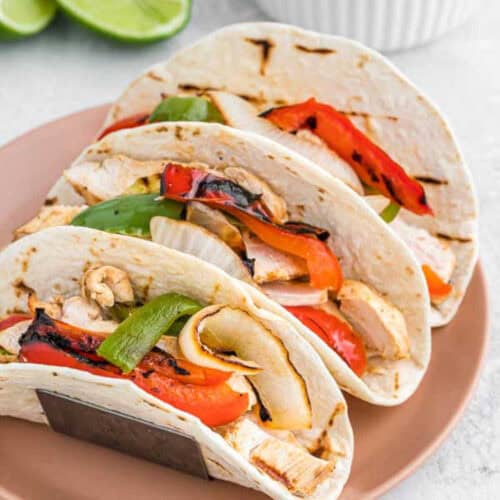 Three sizzling grilled chicken fajitas with vibrant red and green bell peppers and onions, all nestled in flour tortillas on a pink plate. In the background, tantalizing lime slices invite an added zest to these delicious grilled chicken fajitas.