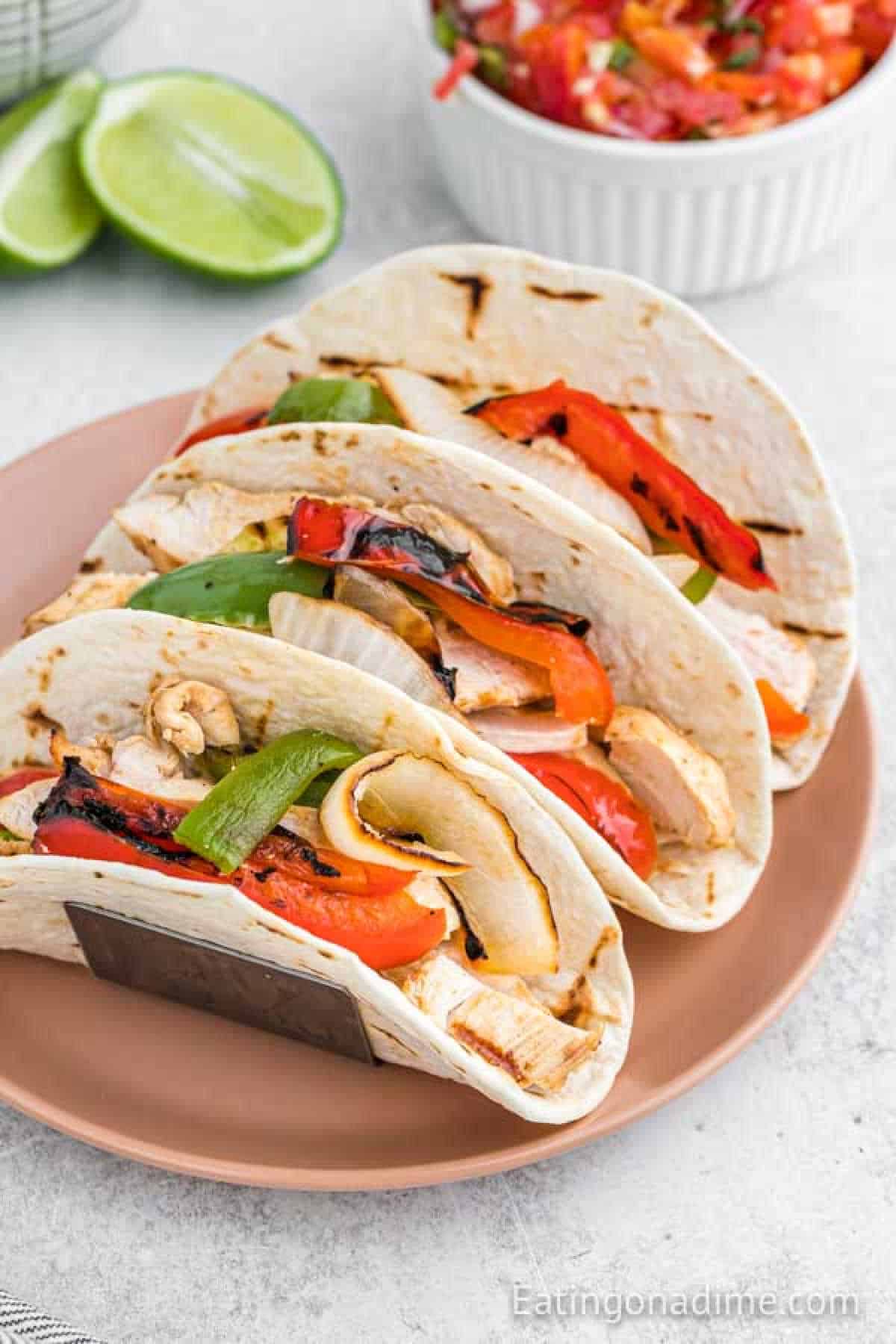 Three grilled chicken fajitas filled with onions and red and green bell peppers rest on a pink plate. In the backdrop, a lime wedge awaits alongside a bowl of salsa, ready to add zest to your meal.