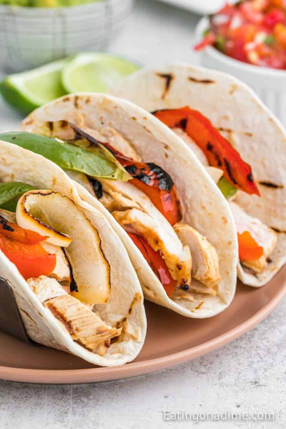 Three grilled chicken fajitas with bell peppers and onions nestle in soft tortillas on a plate. Lime wedges and a bowl of salsa await eagerly in the background, ready to complement the savory delight of these iconic Grilled Chicken Fajitas.