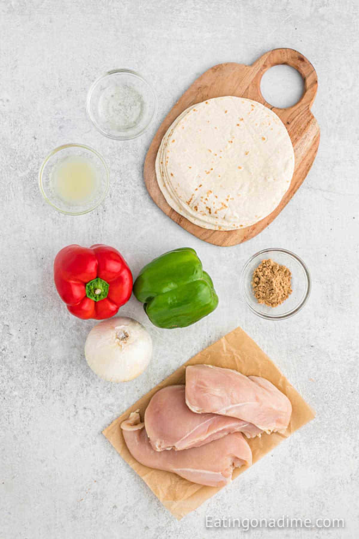 The ingredients for grilled chicken fajitas are artfully arranged: uncooked chicken breasts on paper, vibrant red and green bell peppers, a white onion, a stack of tortillas on a wooden board, a bowl of seasoning, and a small dish with liquid.