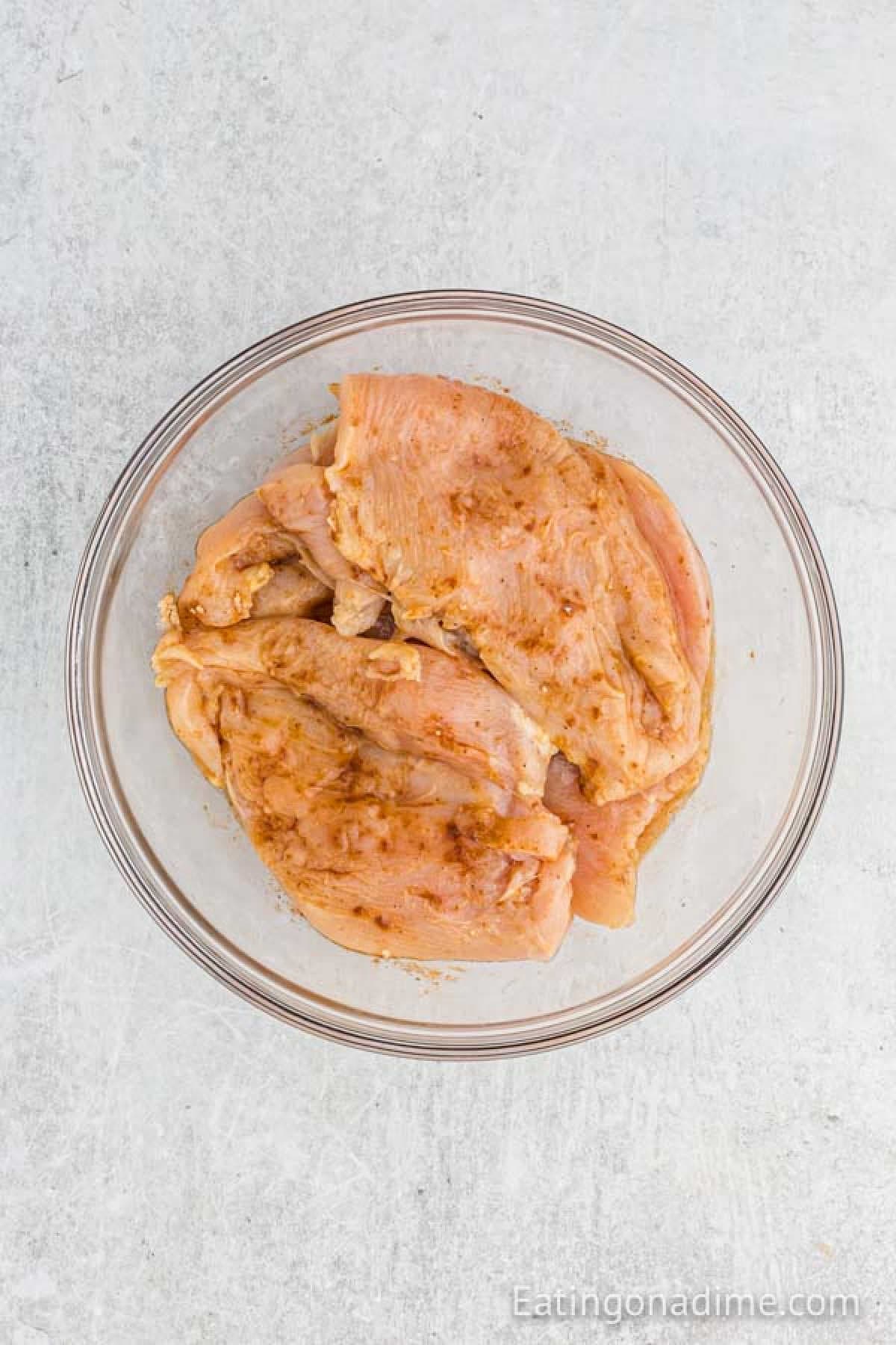 Raw chicken breasts seasoned with spices, perfect for grilled chicken fajitas, are placed in a clear glass bowl on a light gray surface.