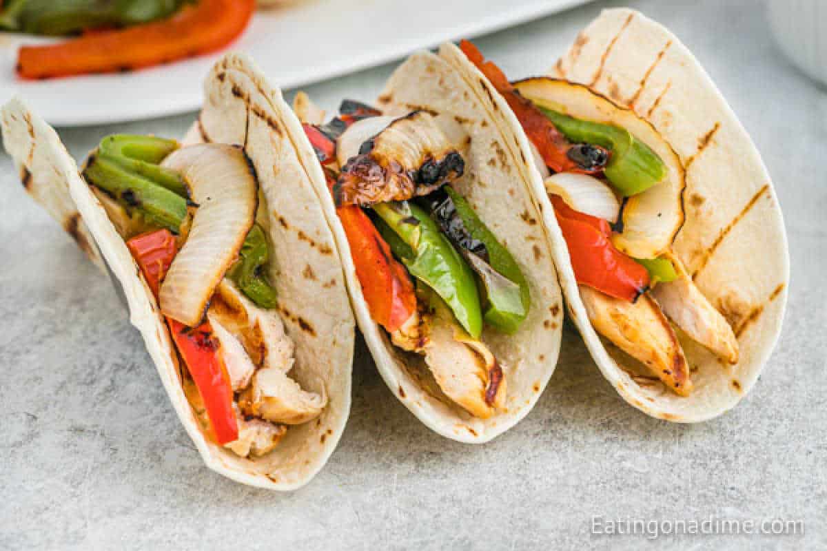 Three succulent grilled chicken fajitas with charred onions and bell peppers nestled in soft tortillas, arranged side by side on a light surface.