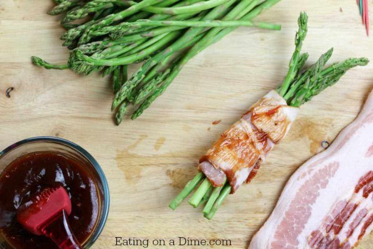 Fresh asparagus spears wrapped in raw bacon rest on a wooden surface alongside a bundle of asparagus and an extra strip of bacon, illustrating a delightful bacon-wrapped asparagus recipe. Nearby, a small bowl with barbecue sauce and a brush awaits the final touch.