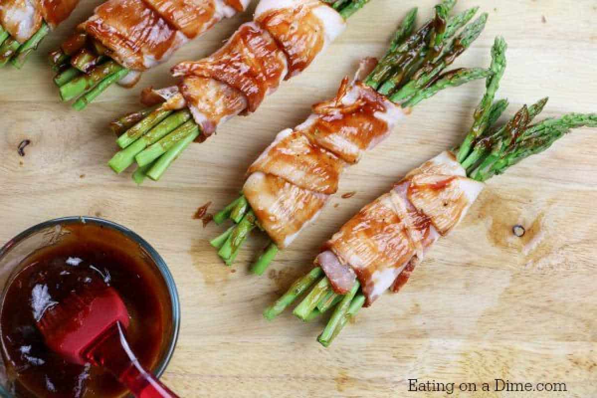 Four bacon-wrapped asparagus bundles are artfully arranged on a wooden surface, partially coated with a rich brown glaze. Nearby, a glass bowl brims with the same glaze and a red basting brush awaits, embodying the essence of this delightful bacon-wrapped asparagus recipe.