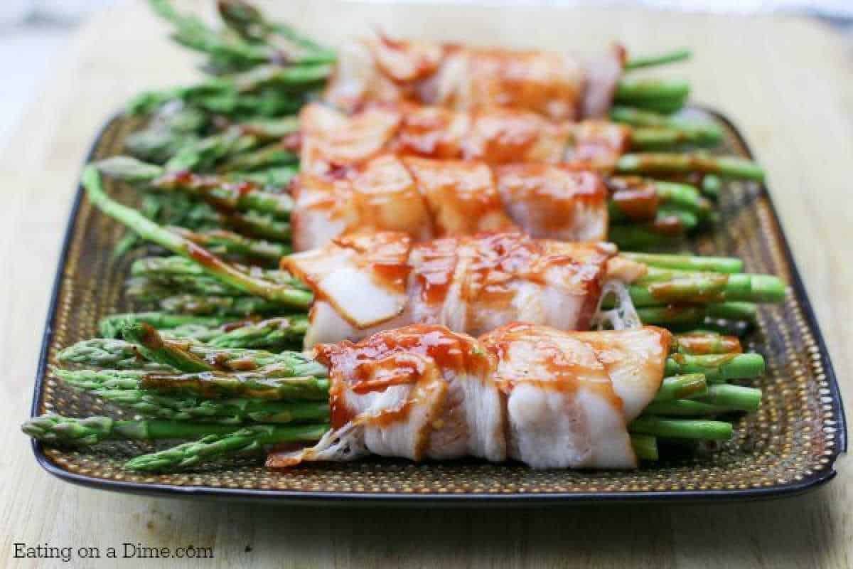 A platter displays neatly arranged bundles of bacon-wrapped asparagus, embodying a delightful recipe. The bundles are topped with a shiny glaze, likely a sauce, giving them an appetizing appearance. The platter rests on a wooden surface.