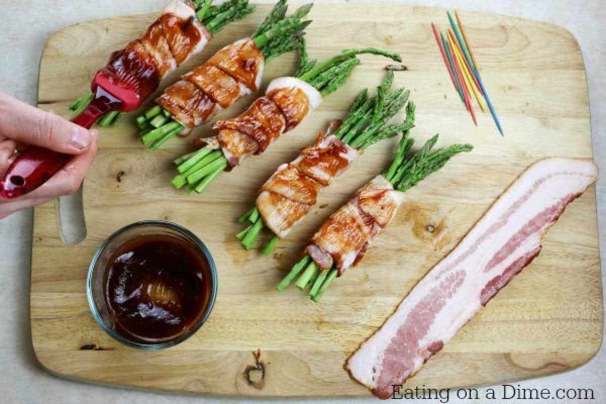 A hand brushes sauce onto bacon-wrapped asparagus bundles on a wooden board, perfecting this delicious bacon-wrapped asparagus recipe. Nearby, a glass bowl of sauce and strips of uncooked bacon await their turn, while colorful toothpicks lie in the corner next to the "Eating on a Dime.com" text.