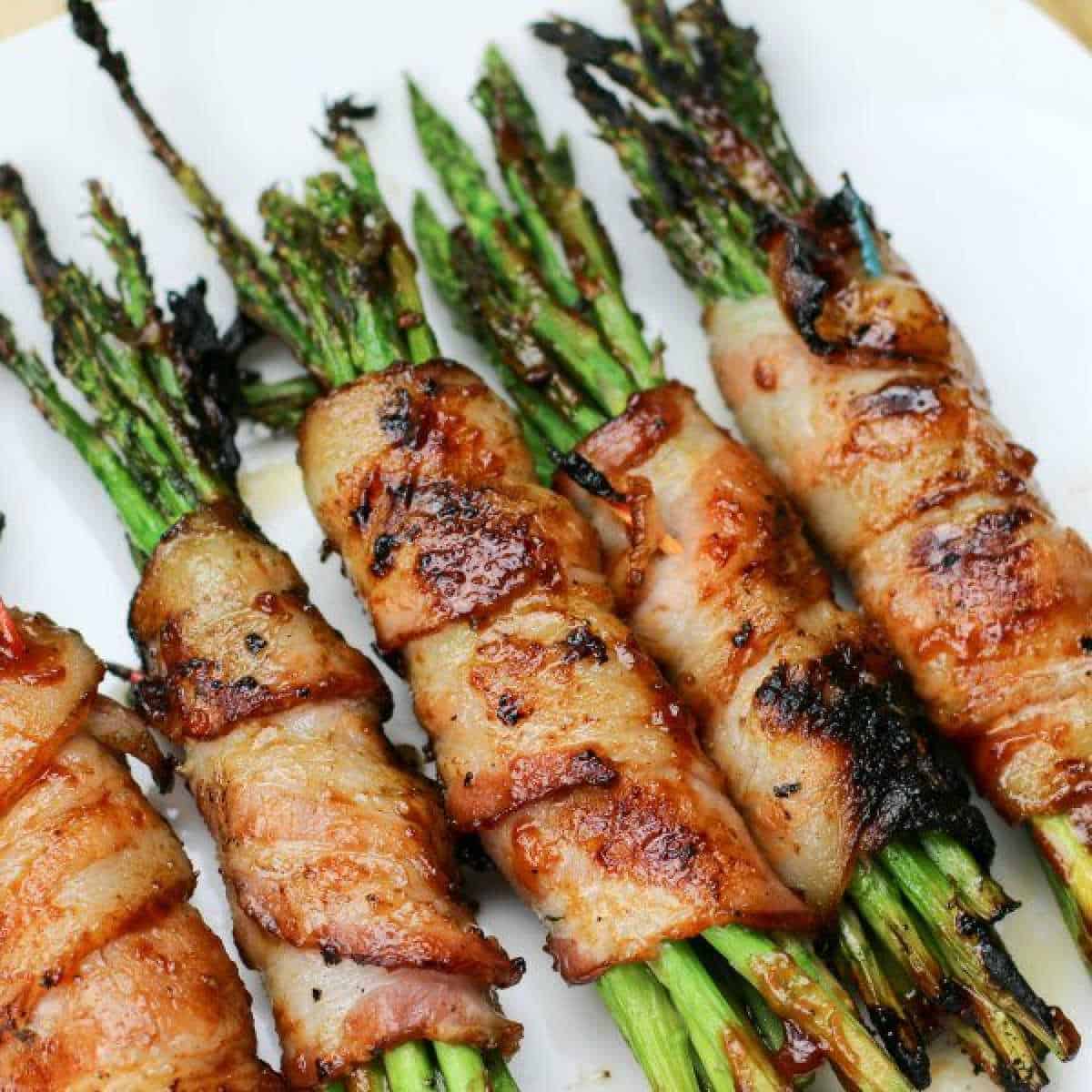 Close-up of a bacon-wrapped asparagus recipe, featuring spears enveloped in crispy, golden-brown bacon. There are four bundles on a white plate, with charred edges hinting at grilling or roasting perfection.