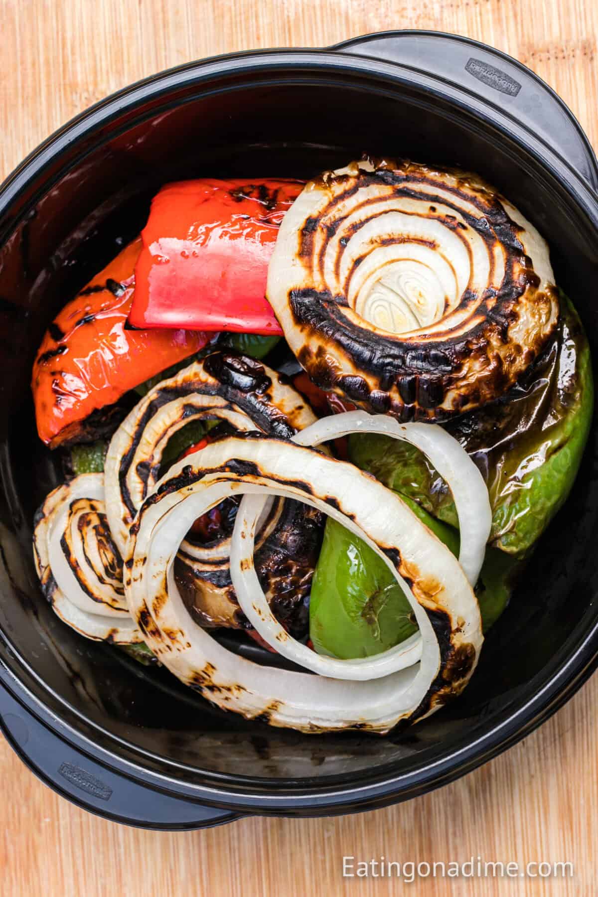 A bowl filled with grilled vegetables, including red and green bell peppers and charred onion rings, is reminiscent of classic grilled chicken fajitas. The colorful veggies are artfully arranged in a round black dish on a wooden surface.