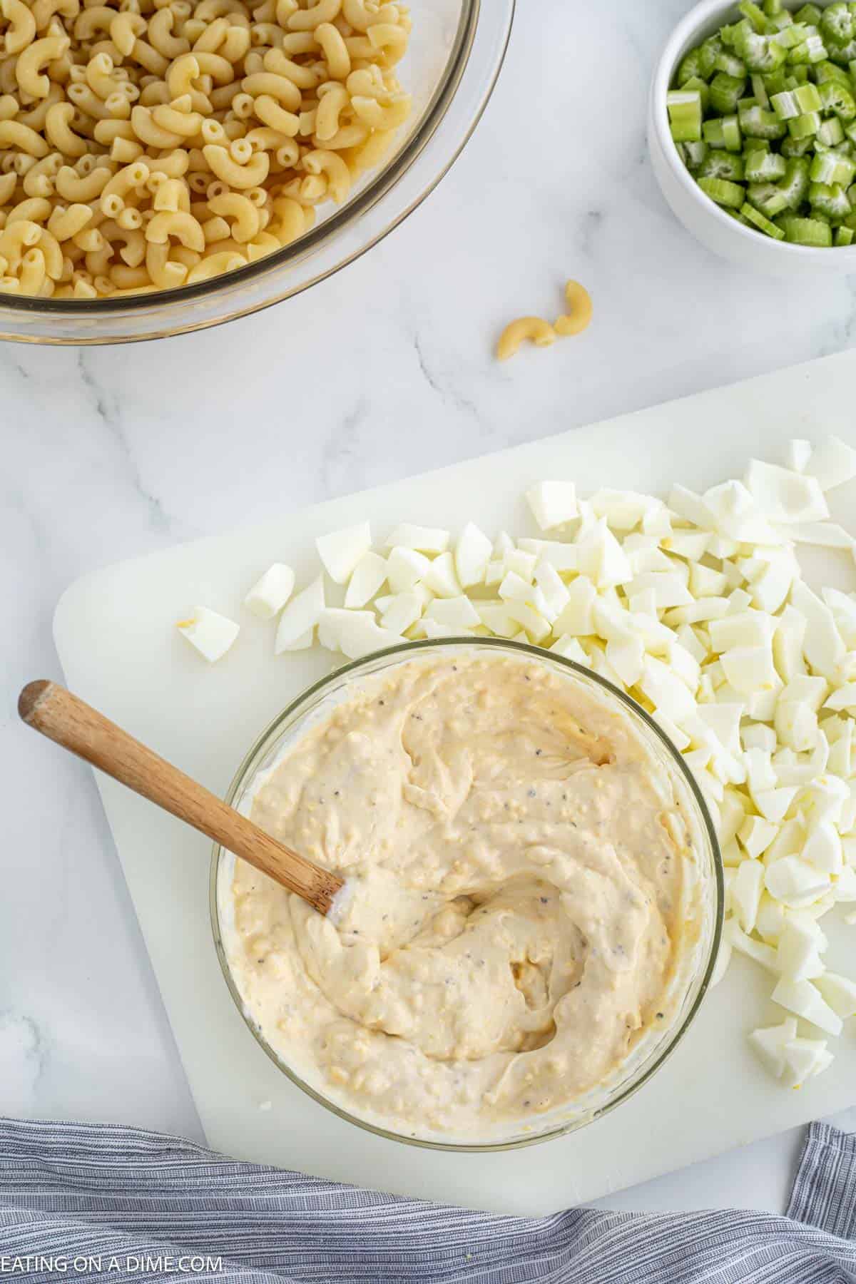 Mayonnaise mixture in a bowl with a wooden spoon with a side of chopped egg whites and a bowl of elbow macaroni noodles