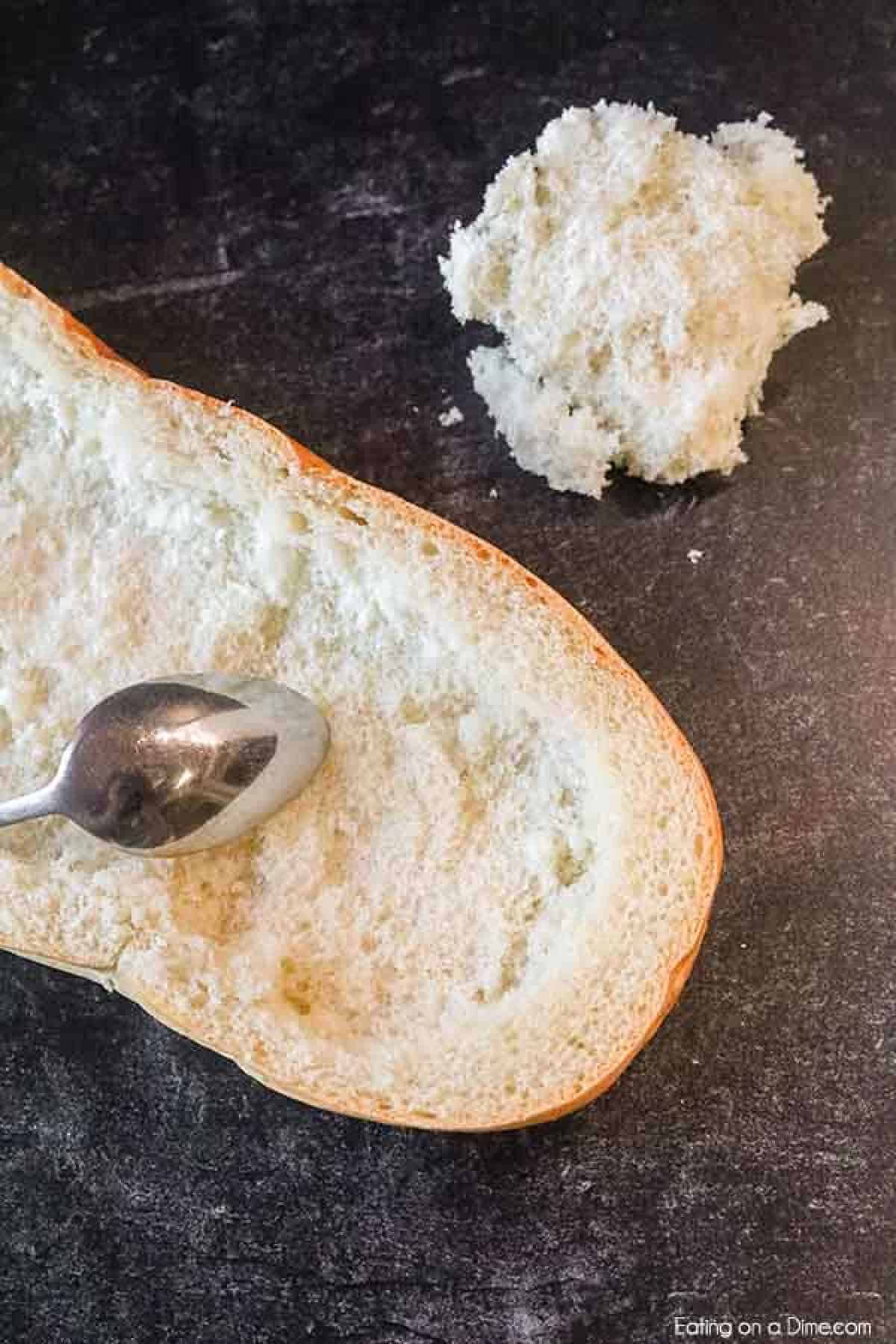 A piece of bread with the soft inner part scooped out using a spoon rests on a dark surface, alongside some removed crumbs, ready to be transformed into a delicious sloppy joes stuffed French bread recipe.