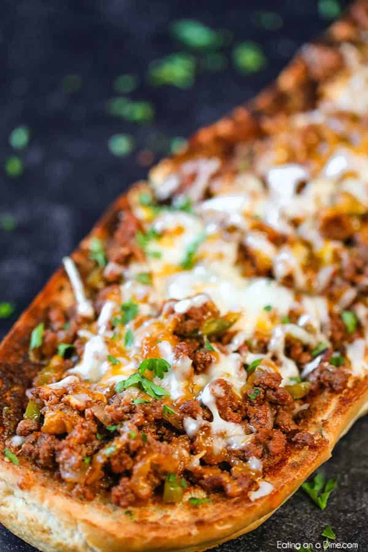 A close-up of a toasted bread topped with melted cheese, seasoned ground meat, and chopped green herbs. This savory dish, reminiscent of a sloppy joes stuffed French bread recipe, is garnished with sprigs of fresh parsley on a dark background.