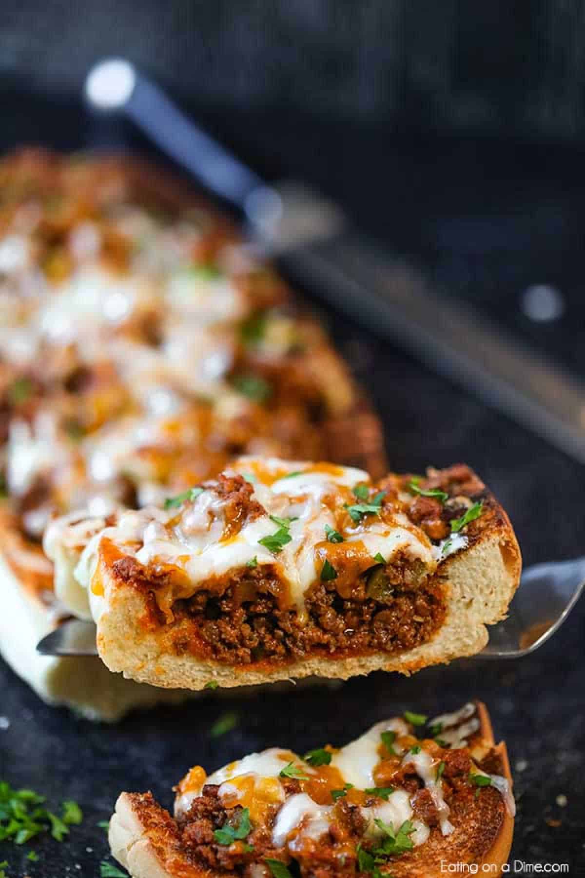 A close-up of a baked stuffed French bread loaf filled with seasoned ground meat, reminiscent of a sloppy joes recipe, and topped with melted cheese. The bread is sliced, and one piece is being lifted to reveal its gooey, cheesy filling. Fresh herbs garnish the top, and a knife is visible in the background.