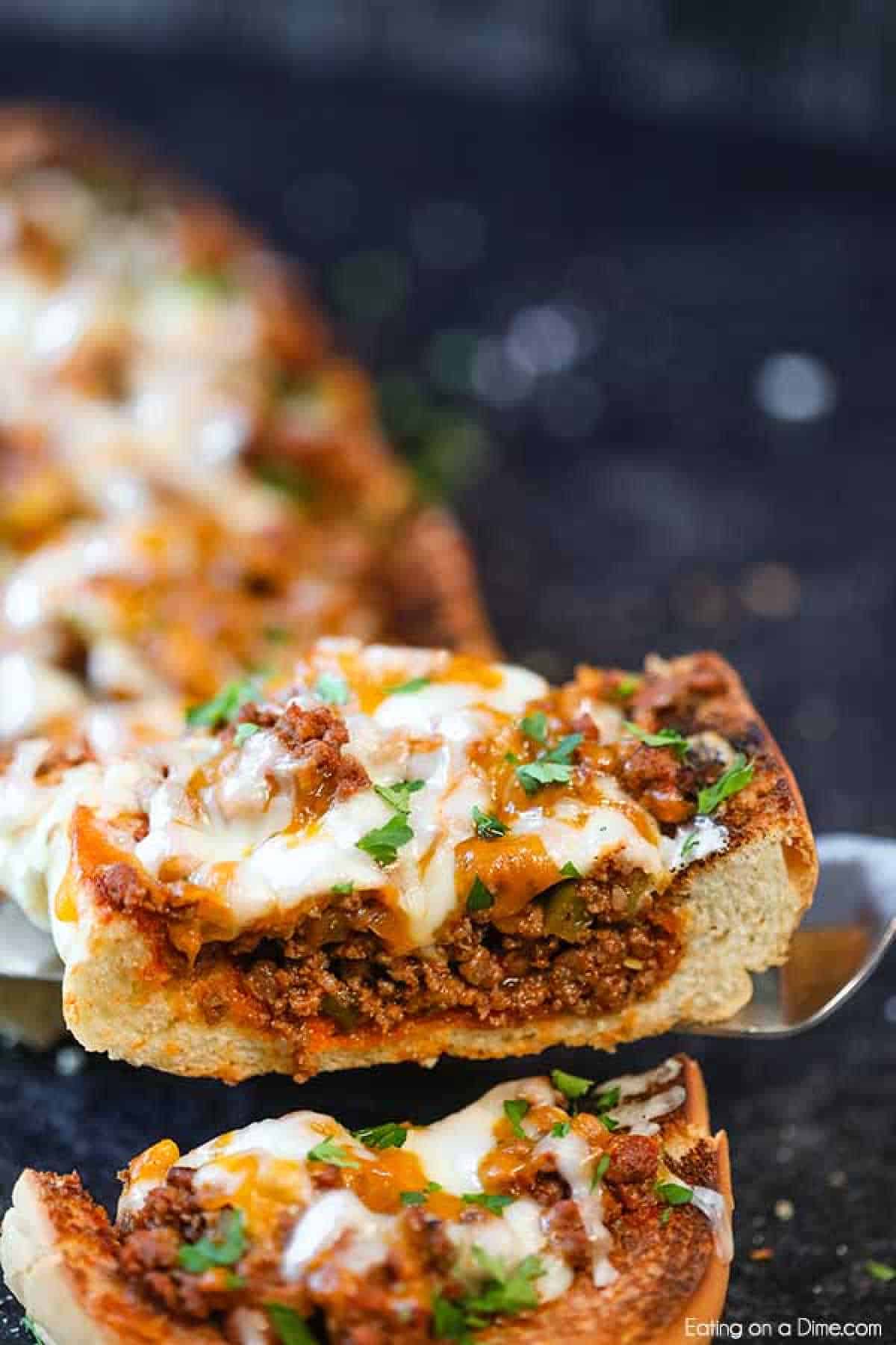 A close-up of cheesy sloppy joes stuffed French bread. The bread is loaded with a savory ground beef mixture, melted cheese, and garnished with chopped parsley. A slice is being lifted with a spatula, revealing the rich filling inside the crusty bread.