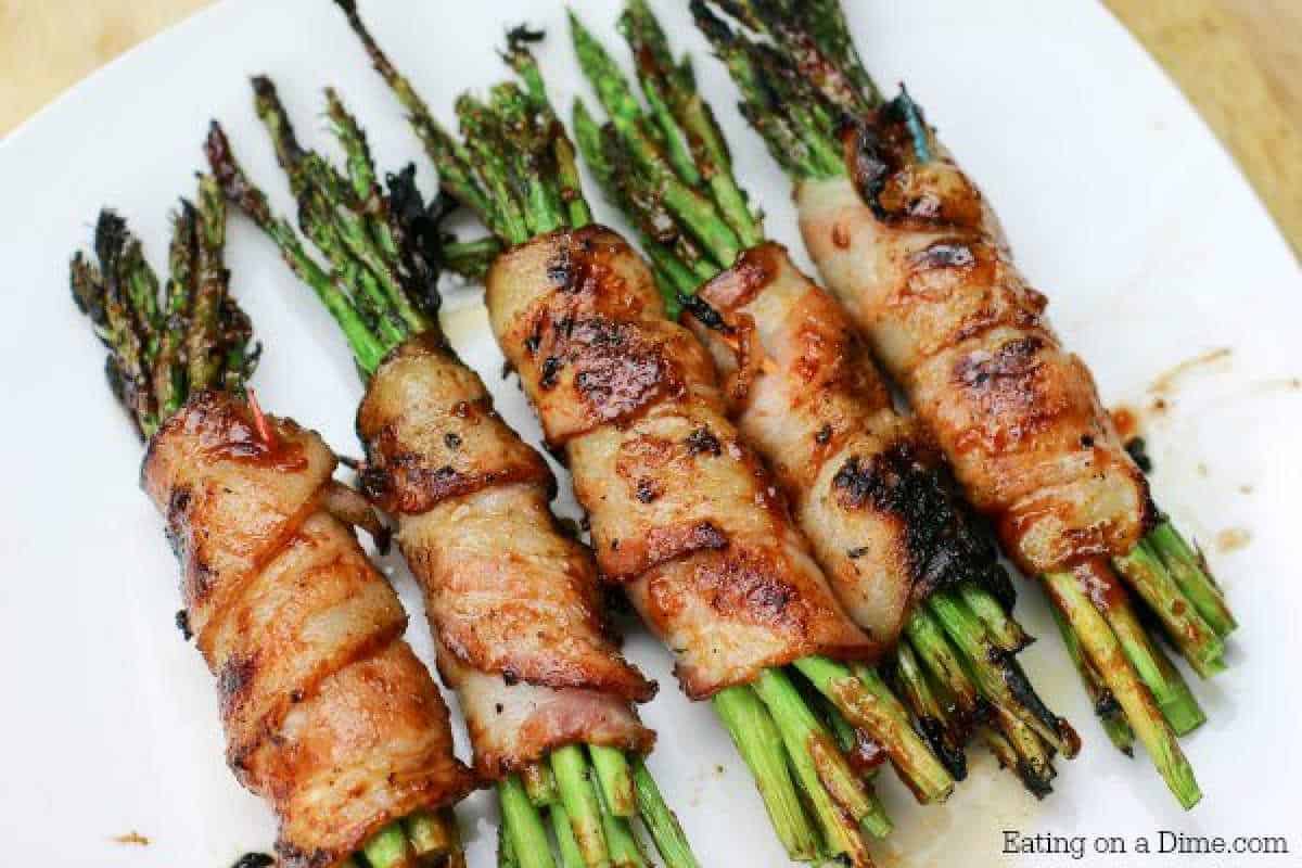 Five bundles of grilled asparagus wrapped in crispy bacon are arranged on a white plate. Each bundle is charred and browned, capturing the essence of a deliciously smoky bacon-wrapped asparagus recipe. A printed watermark reads "Eating on a Dime.com" in the bottom right corner.