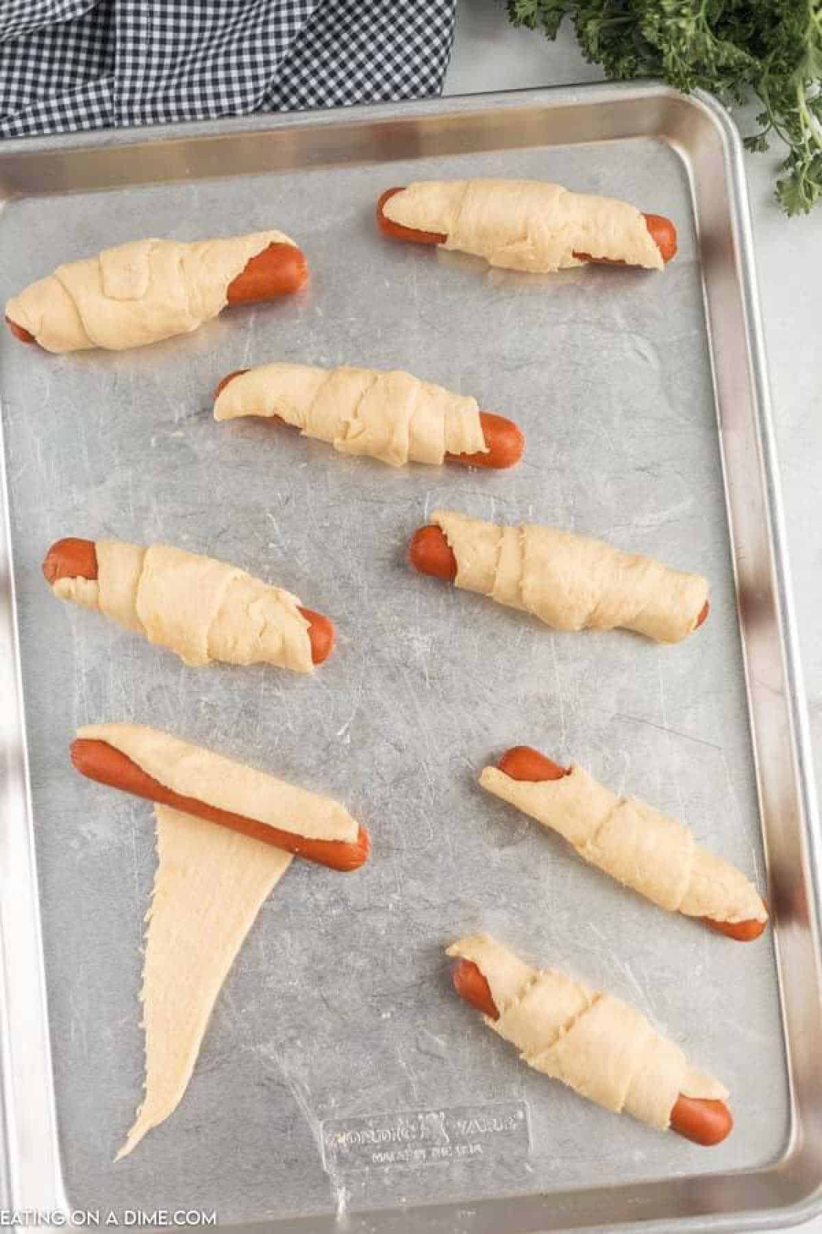 A baking sheet displays eight uncooked crescent roll pigs in a blanket, with hot dogs snugly wrapped in dough. One piece is unwrapped to illustrate the wrapping technique. A checkered cloth and sprigs of parsley provide a charming backdrop for this classic recipe.