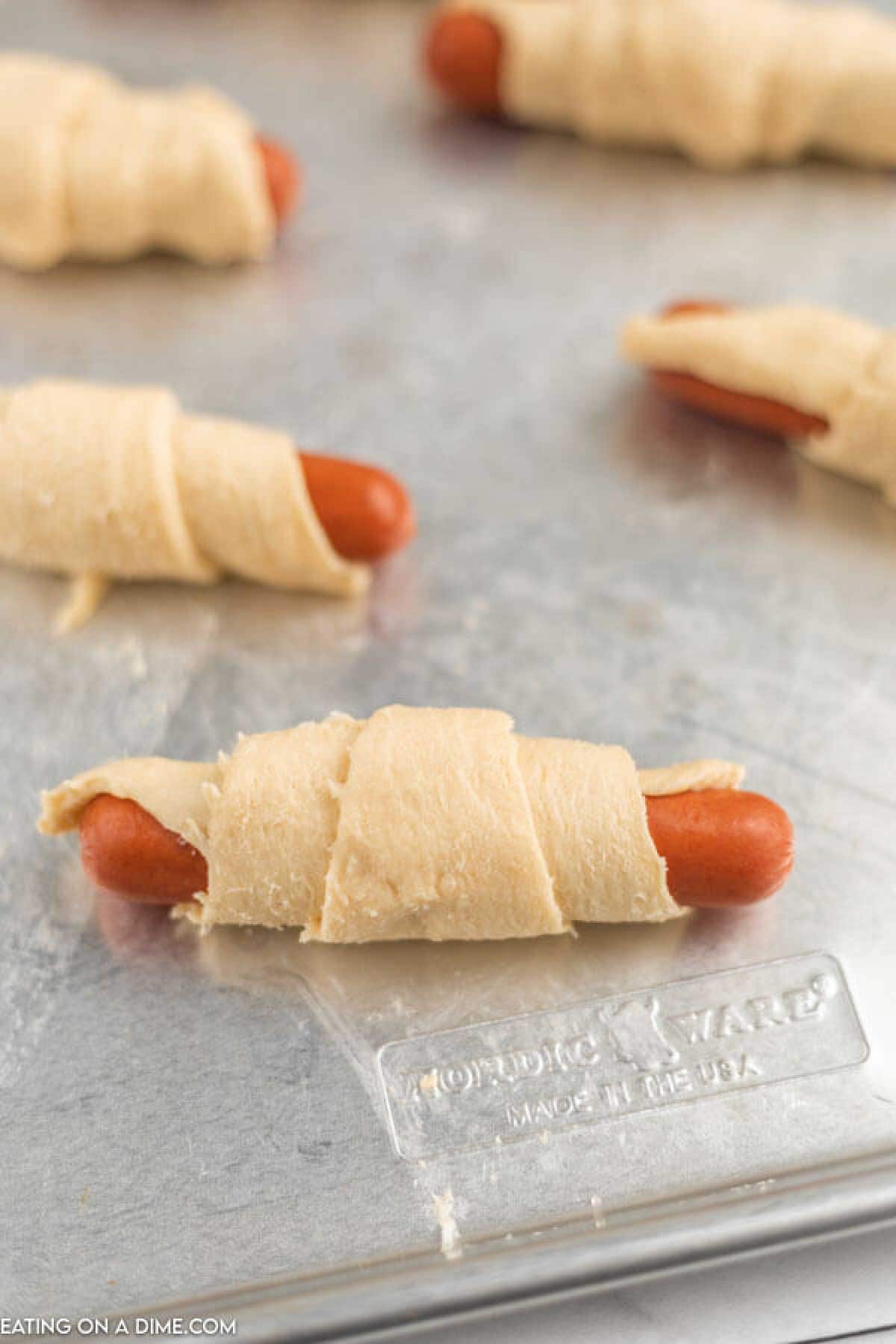 Unbaked crescent roll pigs in a blanket are ready for the oven, with hot dogs wrapped snugly in crescent dough and placed on a metal baking sheet.