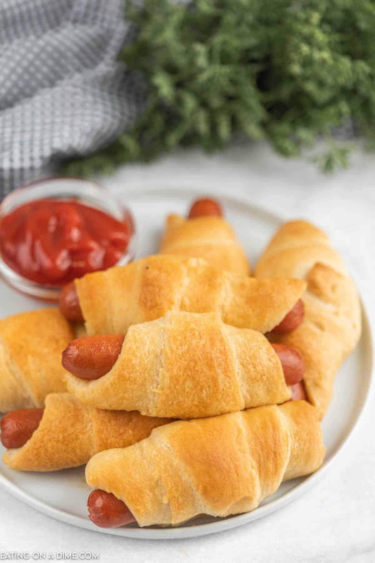 A plate of golden-brown crescent roll pigs in a blanket, served with a small dish of ketchup for dipping, set against a white background. A green plant and a gray cloth add subtle depth to this classic recipe's presentation.