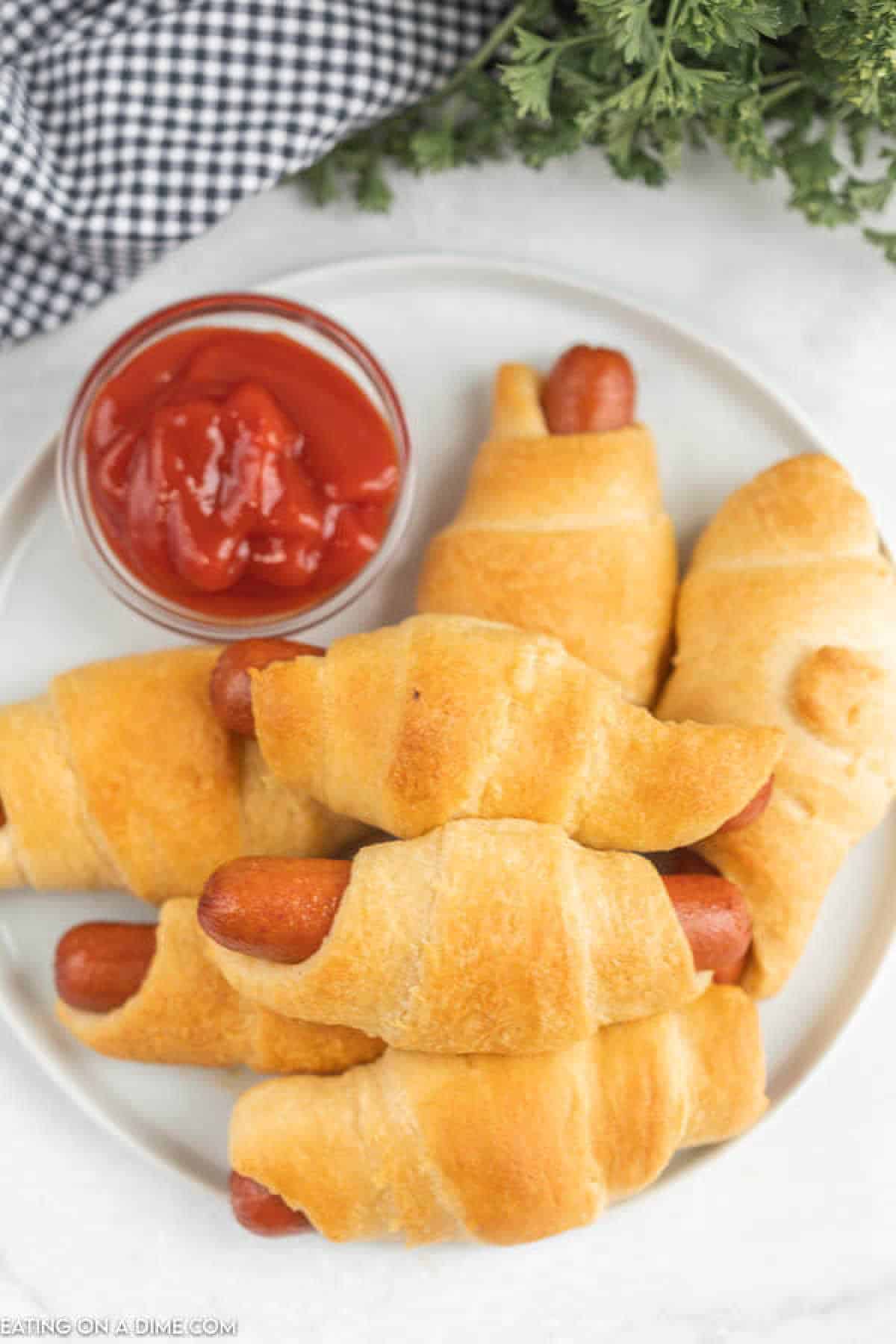 A plate of crescent roll pigs in a blanket, crafted from a classic recipe, is served with a small bowl of ketchup. Fresh parsley and a black-and-white checkered cloth are in the background.