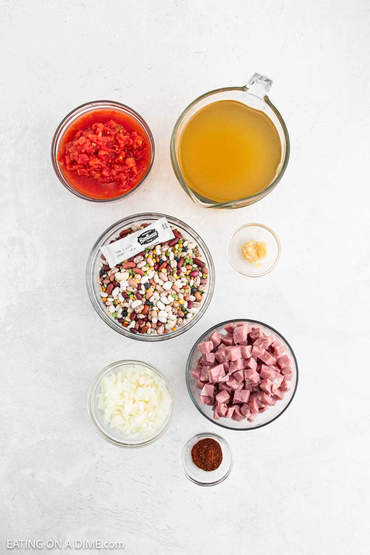 Ingredients for a crock pot 15 bean soup with ham laid out on a white surface: diced tomatoes, broth in a measuring cup, minced garlic in a small bowl, a bag of bean mix, diced onions, cubed ham, and chili powder.