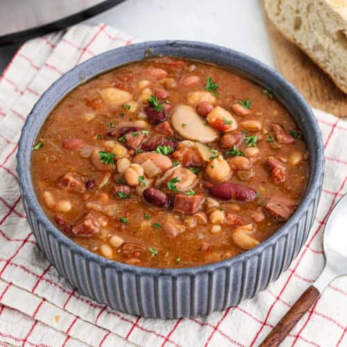 A bowl of hearty crock pot 15 bean soup with ham, garnished with fresh herbs. The stew includes various beans, visible tomato chunks, and pieces of meat. It's placed on a red and white checkered cloth, alongside a slice of bread and a spoon.