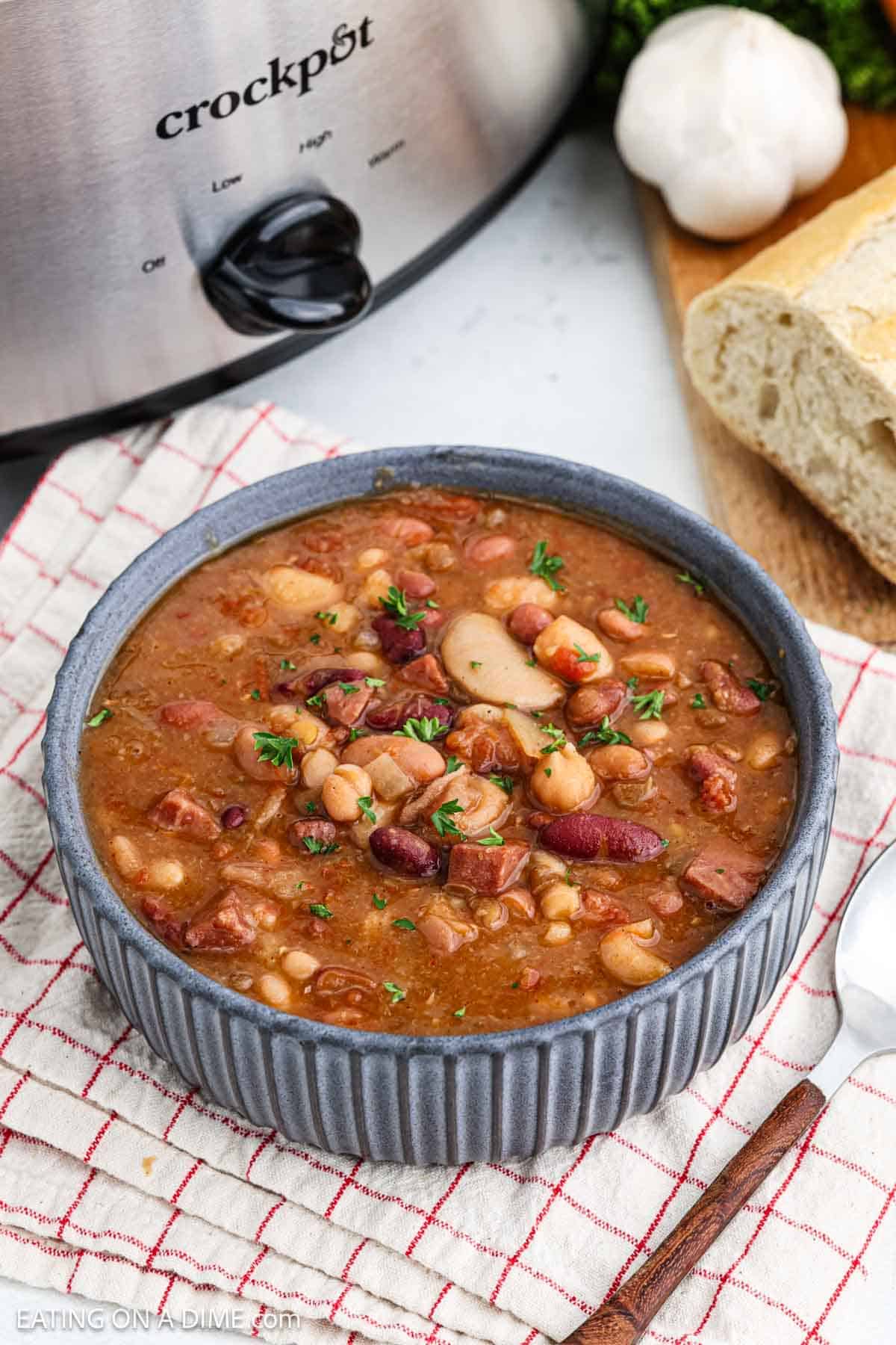 A bowl of hearty 15 bean soup with ham and kidney beans sits on a striped cloth. A crock pot is in the background with a garlic bulb and sliced bread on a cutting board nearby.