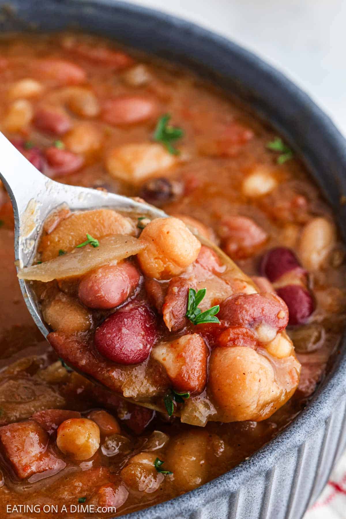 A close-up of a spoonful of crock pot 15 bean stew above a bowl reveals assorted beans and visible herbs within its rich, thick broth. Enhanced with savory ham, the dish appears hearty and flavorful.