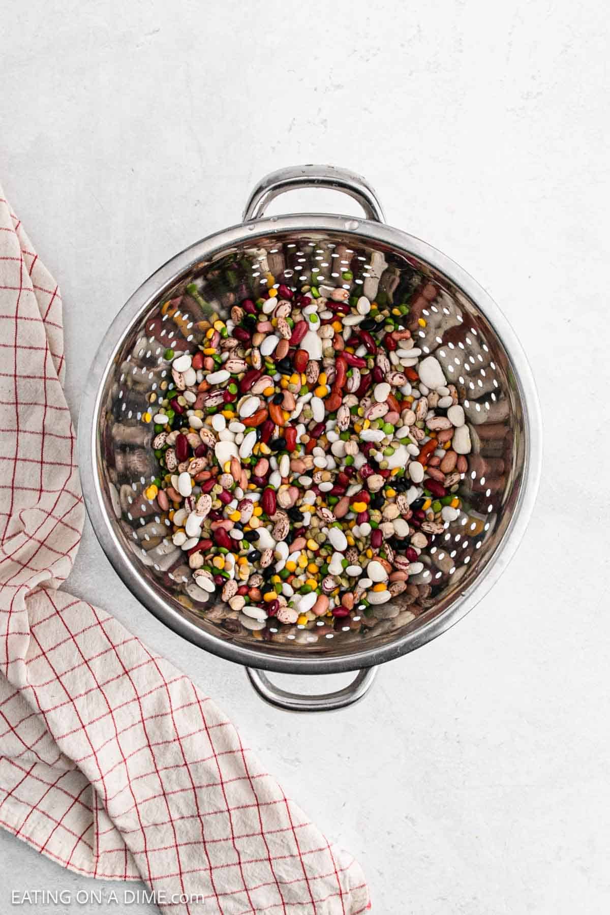 A metal colander filled with a variety of colorful beans, perfect for crock pot 15 bean soup with ham, rests on a light surface. A red and white checkered cloth is crumpled beside the colander.