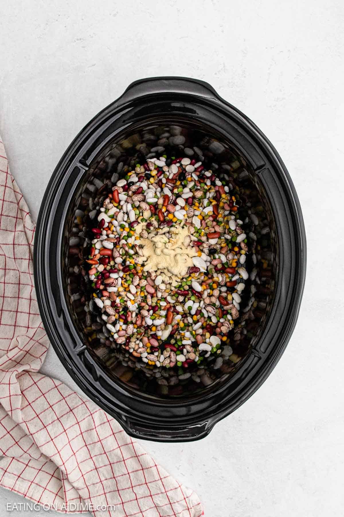 A crock pot filled with 15 bean soup, enriched by diced vegetables, ham, and spices, sits invitingly. A red and white checkered cloth peeks out on the side, resting on a light gray surface.