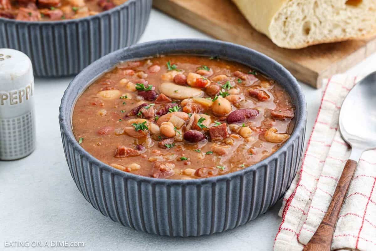 A close-up of a bowl filled with hearty crock pot 15 bean soup with ham, featuring various beans and chunks of meat, garnished with herbs. A spoon rests on a striped cloth beside the bowl, and a loaf of bread is visible in the background.